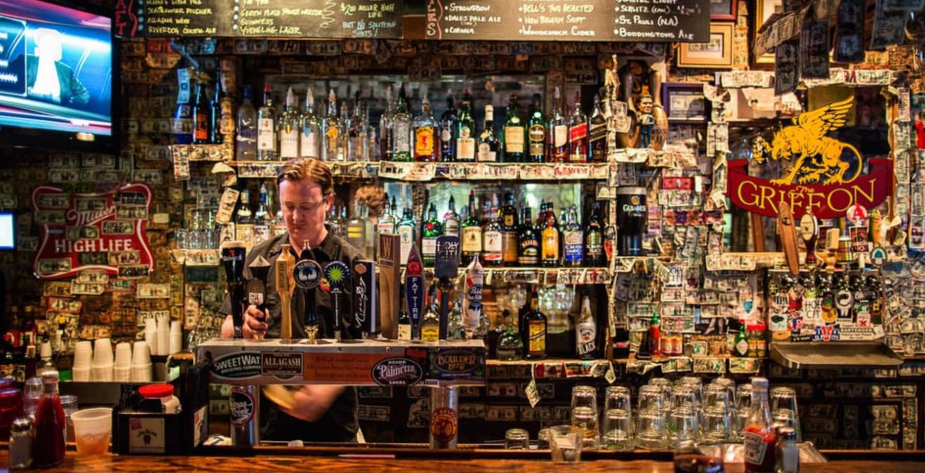 Bartender pouring draft beer at a cozy neighborhood pub — wooden bar with multiple taps, shelves of liquor bottles, a chalkboard menu overhead and walls plastered with dollar bills, stickers and vintage signs.