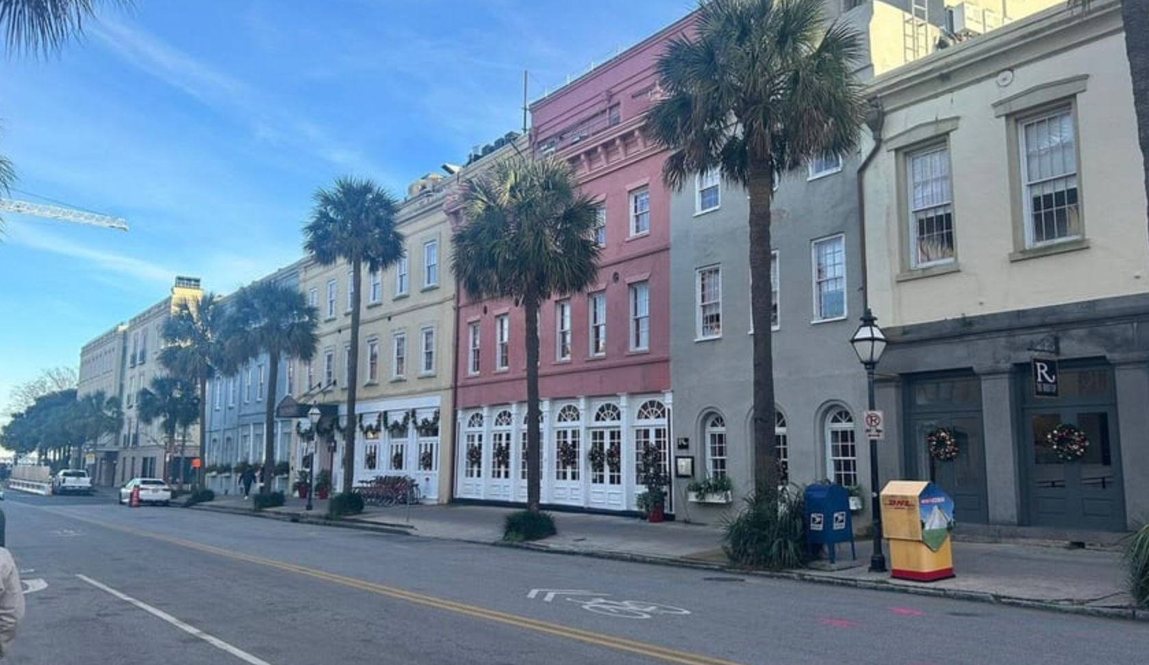 Sunny historic downtown street with pastel rowhouses and tall palm trees lining a quiet coastal road, storefronts decorated with holiday wreaths under a bright blue sky.