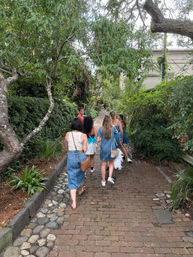 Group of friends walking away along a tree-lined brick pathway with cobblestone edging and dense green foliage — casual summer outfits on a relaxed urban garden stroll.