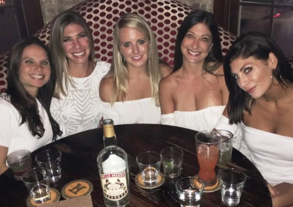 Five friends in white tops smiling in a round booth at a dim cocktail bar, wooden table with mixed drinks, empty glasses, coasters and a bottle for a girls' night out.