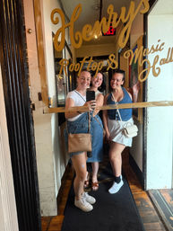 Mirror selfie of three smiling friends in denim and white summer outfits posing in front of a gold-lettered glass entrance advertising a rooftop music hall