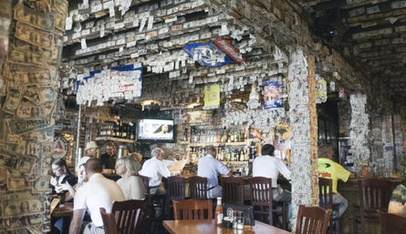 Quirky bar interior with dollar bills plastered on the ceiling and pillars, patrons seated at wooden bar and tables, bottles and a TV behind the bar.