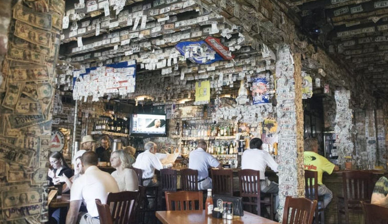 Quirky bar interior with dollar bills plastered on the ceiling and pillars, patrons seated at wooden bar and tables, bottles and a TV behind the bar.