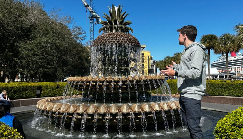 Person gesturing beside the iconic pineapple-shaped fountain at Charleston waterfront park, cascading water tiers, palm trees and a cruise ship under a bright blue sky.