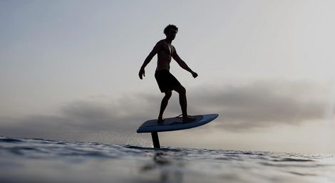 Silhouette of a rider on a hydrofoil surfboard (e-foil) gliding above calm ocean waves at sunset, coastal water sports scene.