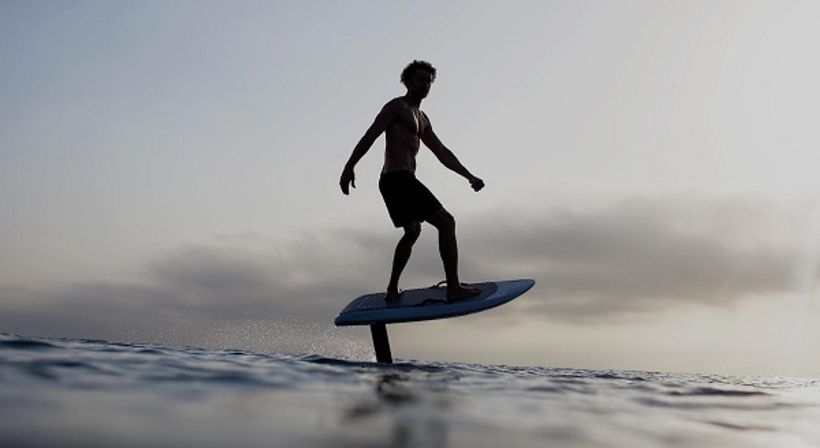 Silhouette of a rider on a hydrofoil surfboard (e-foil) gliding above calm ocean waves at sunset, coastal water sports scene.