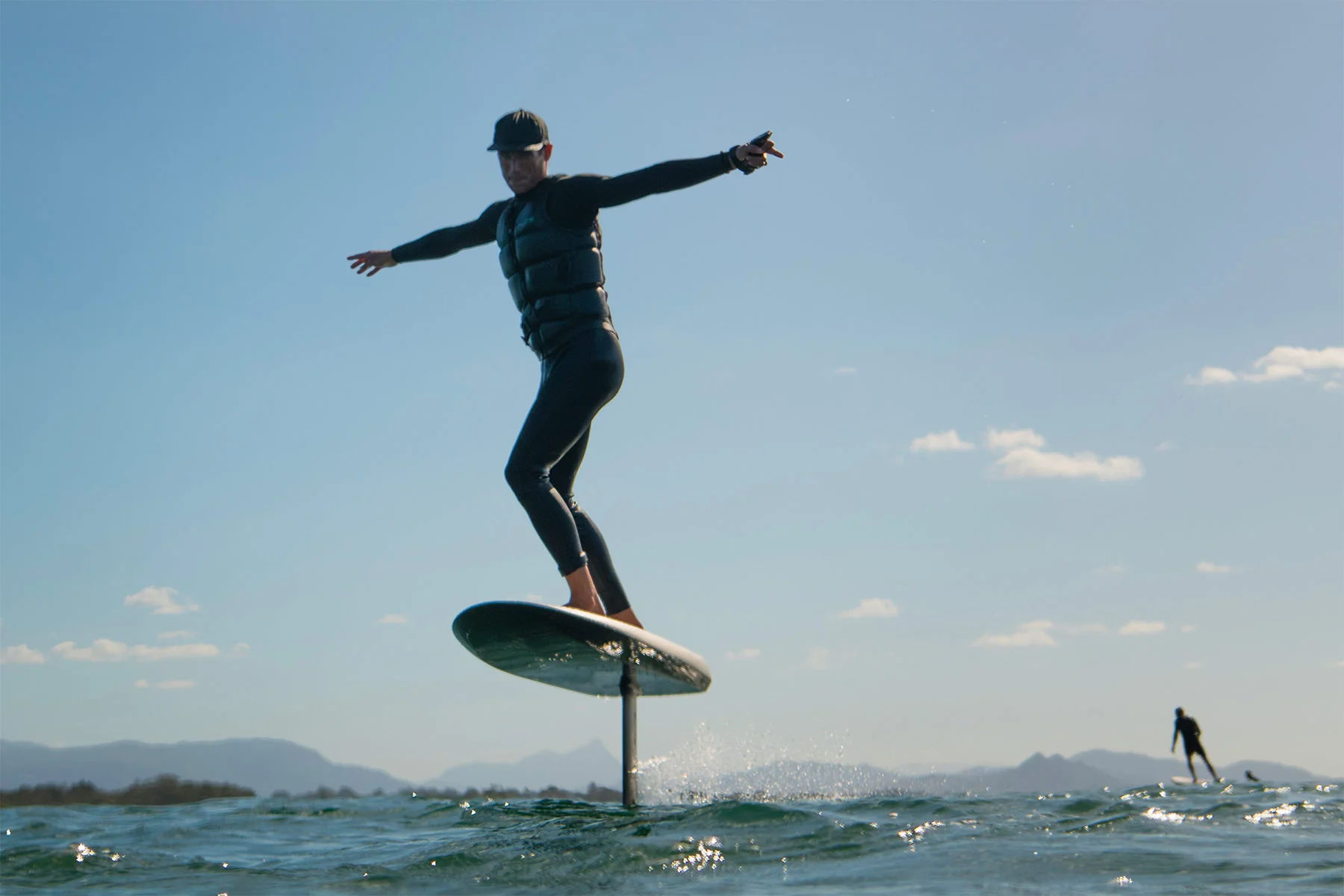Person in wetsuit and life vest balancing on a hydrofoil surfboard, gliding above choppy coastal water under a clear blue sky with distant mountains and another rider in the background.