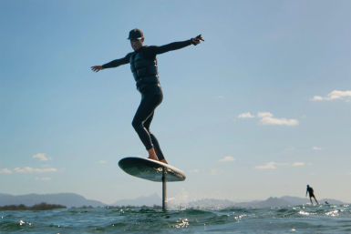 Person in wetsuit and life vest balancing on a hydrofoil surfboard, gliding above choppy coastal water under a clear blue sky with distant mountains and another rider in the background.