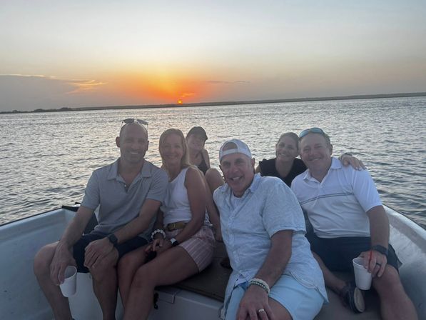 Group of six adults smiling on a small boat at sunset over calm coastal waters, orange sun dipping on the horizon, casual summer outfits and drinks