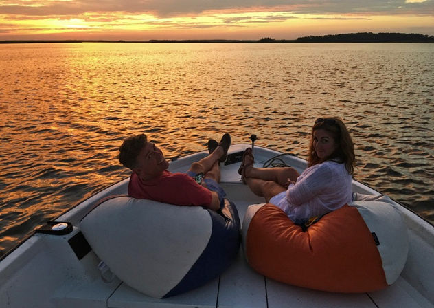 Two people lounging on orange and blue beanbags at the bow of a small boat, watching a golden sunset over calm lake waters and a distant tree-lined horizon.