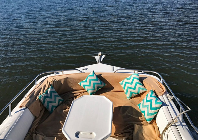 Aerial view of a boat bow lounge: sunlit tan cushions, white table and four turquoise chevron throw pillows overlooking calm open water