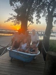 Playful group of six friends sitting in a weathered blue clawfoot tub on a wooden dock at a marina, silhouetted by a golden sunset over the waterfront with boats and oak trees.