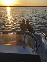 Two friends sitting on a motorboat at golden sunset, sun reflecting across rippling water as a small group relaxes on an evening cruise.