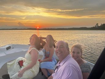 Four adults smiling on a small boat during a golden sunset over a calm bay, distant tree-lined shore, casual summer clothes and a red cup.