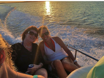 Two smiling women in sunglasses relaxing on a boat at sunset, golden light reflecting on open water and a foamy wake behind them.