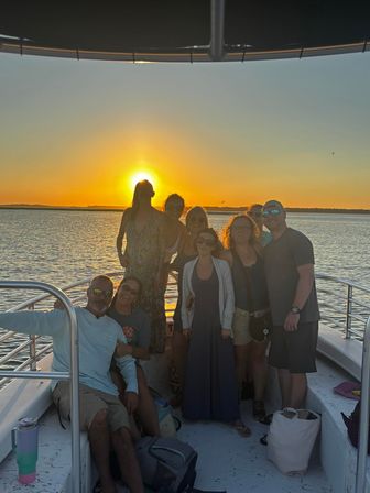 Group of people on a boat deck at golden sunset over a calm coastal bay, relaxed summer outing