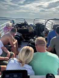 Passengers and packed luggage on a small ferry crossing calm coastal waters with sunlight glinting on the bay.
