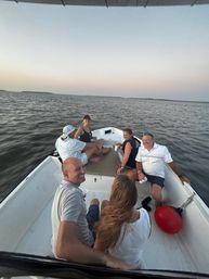 Group of friends relaxing on a small motorboat at dusk, holding drinks and smiling while cruising on calm coastal waters under a pale sunset sky with a red buoy in the boat