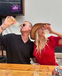 Couple at an outdoor patio beer garden cheering and drinking from bottles — woman in a red dress and straw hat, man in black shirt and sunglasses at a wooden table with plastic cups.