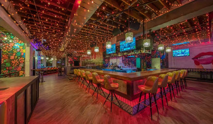 Vibrant downtown cocktail bar interior with a central illuminated wooden square bar, yellow retro bar stools, hanging pendant lamps, twinkling ceiling string lights, neon murals and wall-mounted TVs.