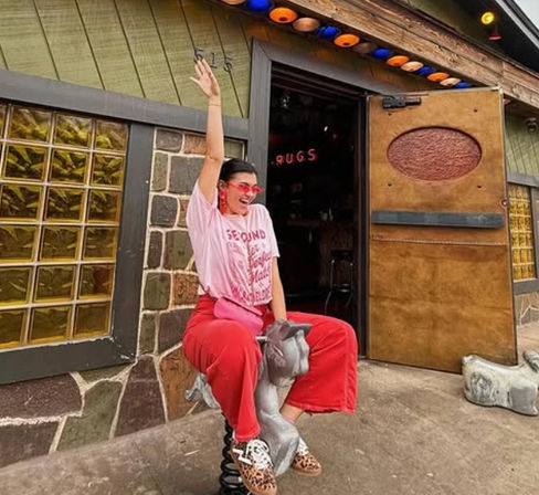 Person in pink t‑shirt and red pants laughing while sitting on a spring-mounted animal ride outside a retro storefront with glass block windows, colorful marquee lights, and an open wooden door.