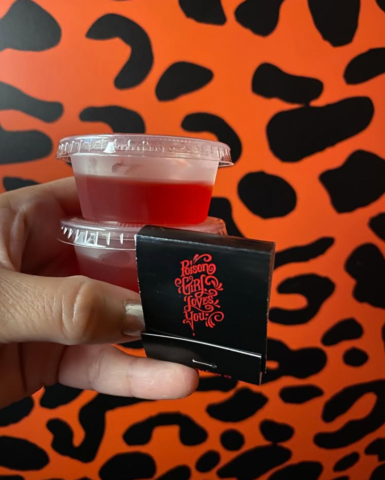 Hand holding two stacked small red gelatin cups with clear lids and a black matchbook featuring red decorative script, set against an orange and black leopard-print wall.
