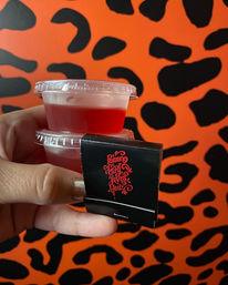 Hand holding two stacked small red gelatin cups with clear lids and a black matchbook featuring red decorative script, set against an orange and black leopard-print wall.