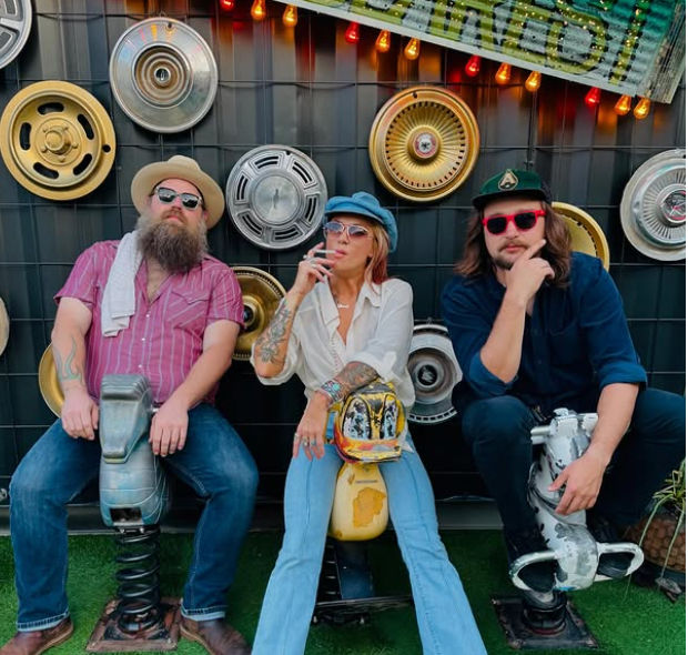 Three people seated on colorful retro ride-on sculptures in front of an outdoor art wall of vintage hubcaps and string lights, wearing hats, sunglasses, and visible tattoos.