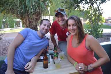 Three smiling friends gathered at an outdoor patio table with drinks, a palm tree and desert landscaping — casual summer hangout.