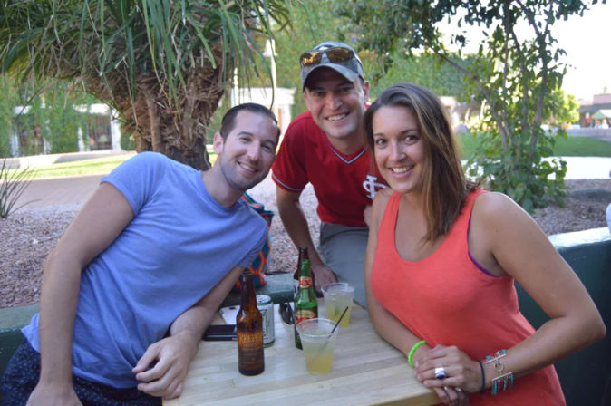 Three smiling friends gathered at an outdoor patio table with drinks, a palm tree and desert landscaping — casual summer hangout.