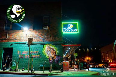 Downtown nighttime scene: neon-lit brick bar entrance with glowing green duck sign, colorful mural reading "It ain't over 'til it's over", parked electric scooters along the curb, and people gathered under string lights.