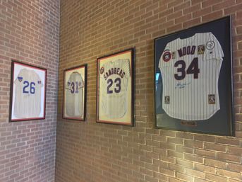 Sports-memorabilia corner with four framed pinstripe baseball jerseys numbered 26, 31, 23 and 34 mounted on a red brick interior wall.