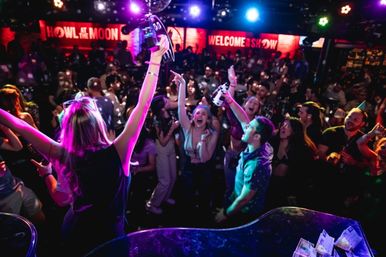 Lively nightclub dance floor filled with a cheering crowd under colorful stage lights and a disco ball, partygoers dancing and waving tambourines and drinks in a high-energy live-show atmosphere.