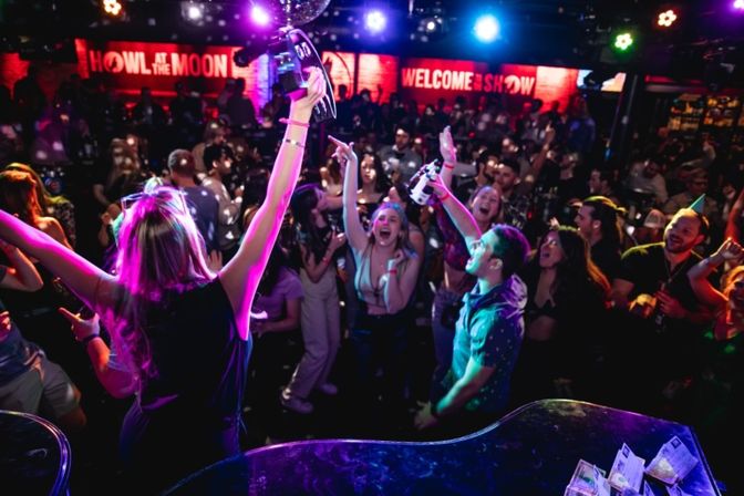 Lively nightclub dance floor filled with a cheering crowd under colorful stage lights and a disco ball, partygoers dancing and waving tambourines and drinks in a high-energy live-show atmosphere.