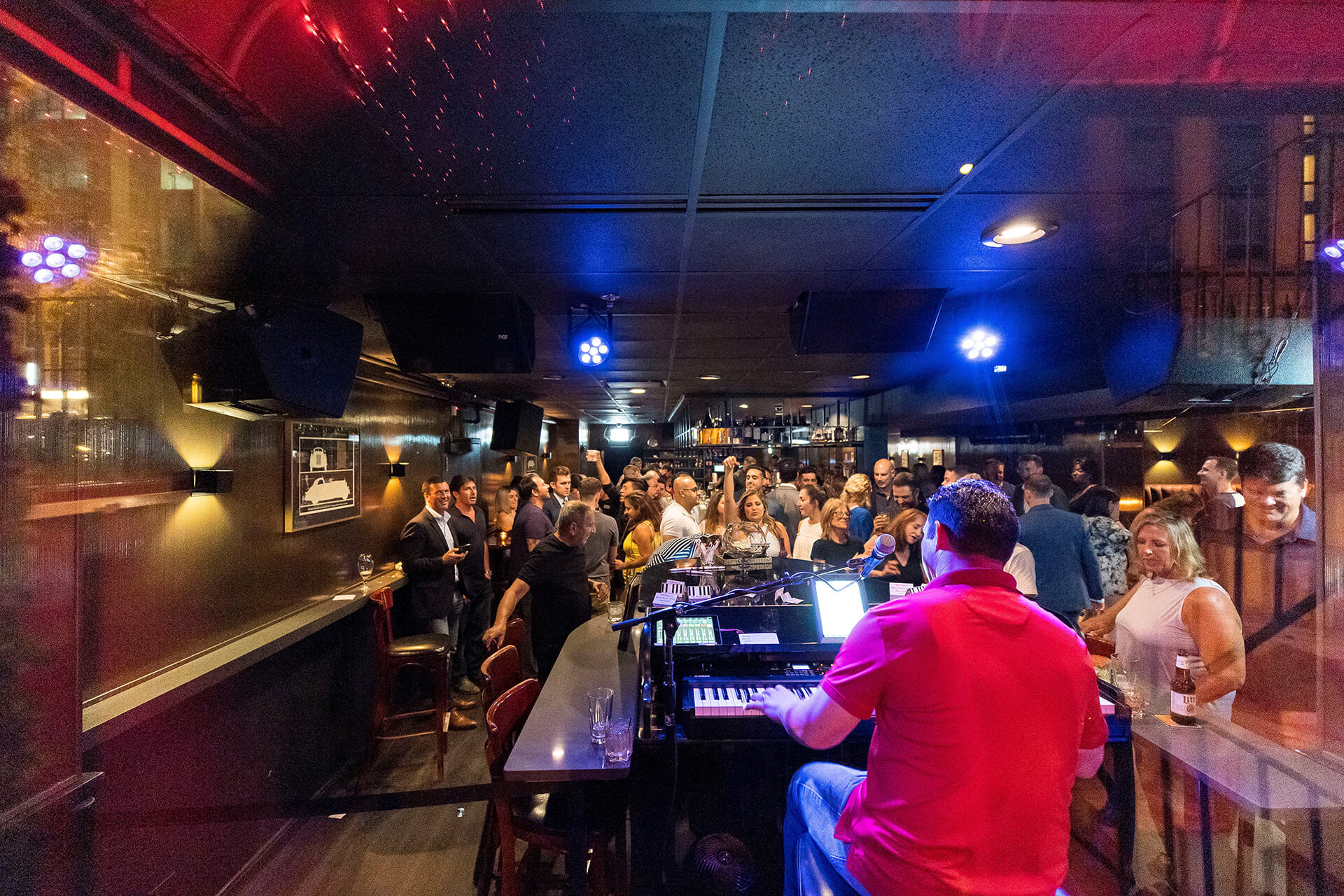 Lively urban piano bar at night with a keyboard player performing under blue lights while a packed crowd of patrons holds drinks and dances.