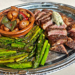 Juicy sliced medium-rare steak topped with herb butter, charred asparagus spears and roasted baby potatoes with peppers on a decorative silver platter.