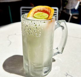Icy frozen margarita in a frosted mug with a salted-herb rim, garnished with a grapefruit wedge and lime slice, sitting on a sunlit outdoor patio table.
