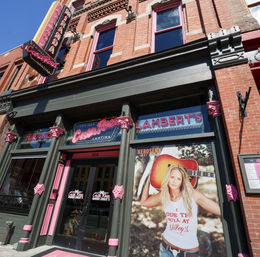 Sunlit red-brick downtown storefront with pink-trimmed columns, neon signage and a large poster of a woman holding an acoustic guitar at the entrance to a Tex-Mex cantina.