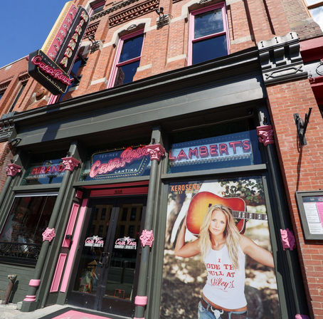 Sunlit red-brick downtown storefront with pink-trimmed columns, neon signage and a large poster of a woman holding an acoustic guitar at the entrance to a Tex-Mex cantina.