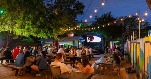 Evening outdoor beer garden patio with string lights, communal picnic tables and groups watching a large screen — casual drinks and social gathering