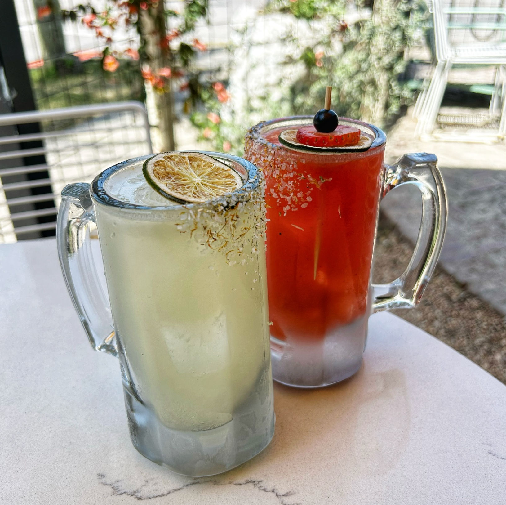 Two frosty cocktail mugs on an outdoor patio table — a pale citrus drink with a dried lime slice and salted rim, and a bright red fruit cocktail garnished with a strawberry slice and cherry.