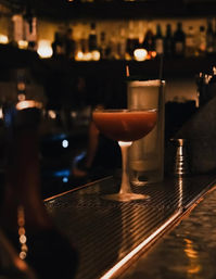 Reddish cocktail in a coupe glass on a dimly lit bar counter, with a frosty highball and blurred bottles and candlelight in the warm background – cozy speakeasy vibe.