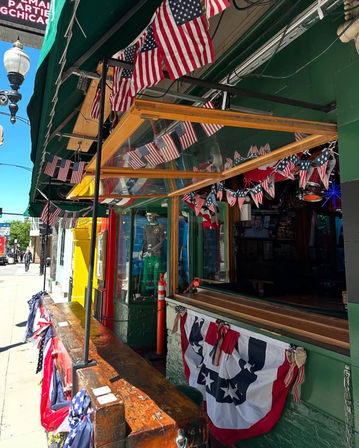 Downtown sidewalk bar with open serving window, wooden outdoor counter and rows of American flags and patriotic bunting under a green awning