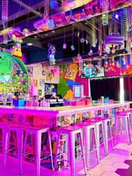 Vibrant neon bar interior with pink and purple lighting, long white cocktail counter and metal bar stools, retro pop-culture collage on the bar faces, hanging slinky coils, CDs and plush toys from the ceiling, and a colorful back-bar display.