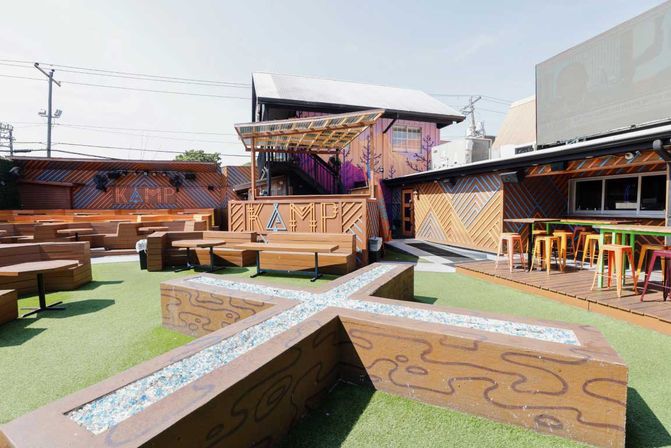 Sunny urban rooftop beer garden with wooden bench seating, colorful bar stools at a raised bar, geometric wooden planters filled with decorative crushed glass, artificial turf, patterned wood facades and a small covered stage.