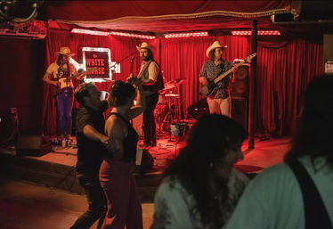 Couples dancing on a small dance floor as a three-piece country band in cowboy hats performs on a red-curtained stage in an intimate live music bar under warm red lighting.