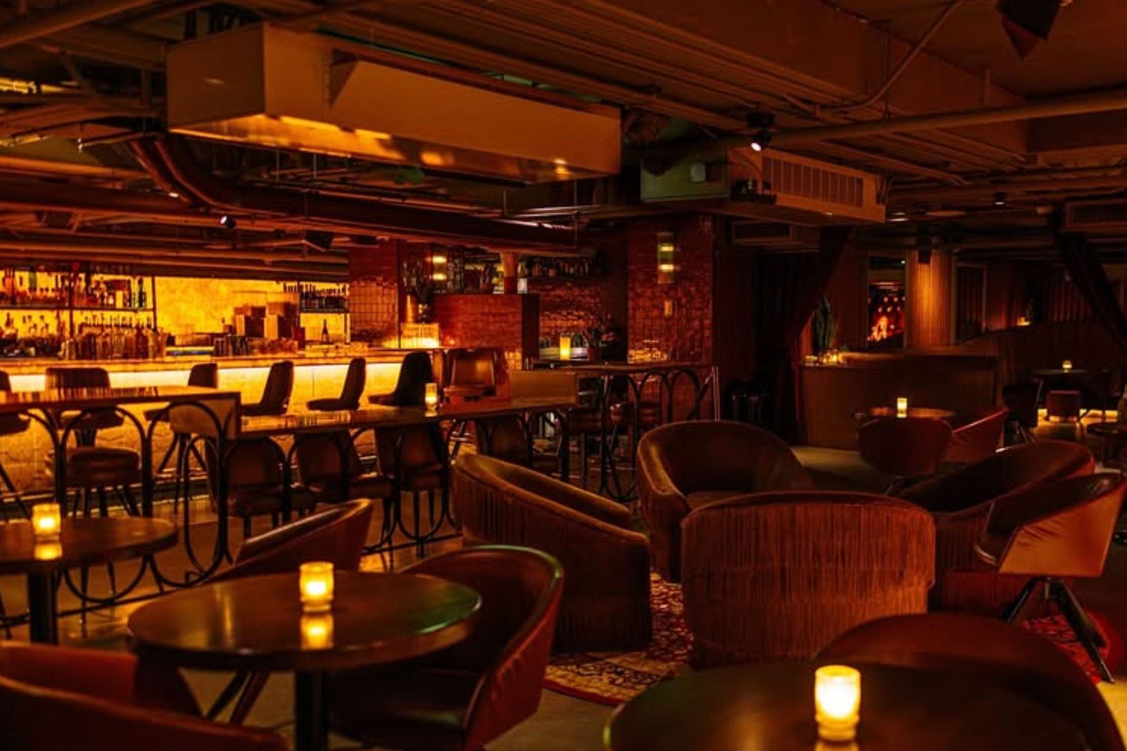 Candlelit underground cocktail lounge with velvet armchairs and round tables facing a glowing backlit bar under warm amber lighting, cozy intimate evening vibe.
