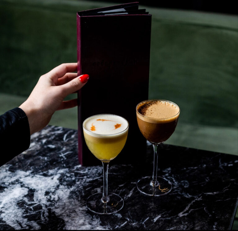 Two cocktails in coupe glasses on a black marble table at a cocktail bar — a frothy yellow drink with orange garnish and a coffee-colored espresso-style cocktail, with a hand with red nails holding a dark menu against a green velvet banquette