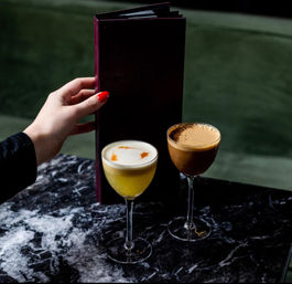 Two cocktails in coupe glasses on a black marble table at a cocktail bar — a frothy yellow drink with orange garnish and a coffee-colored espresso-style cocktail, with a hand with red nails holding a dark menu against a green velvet banquette