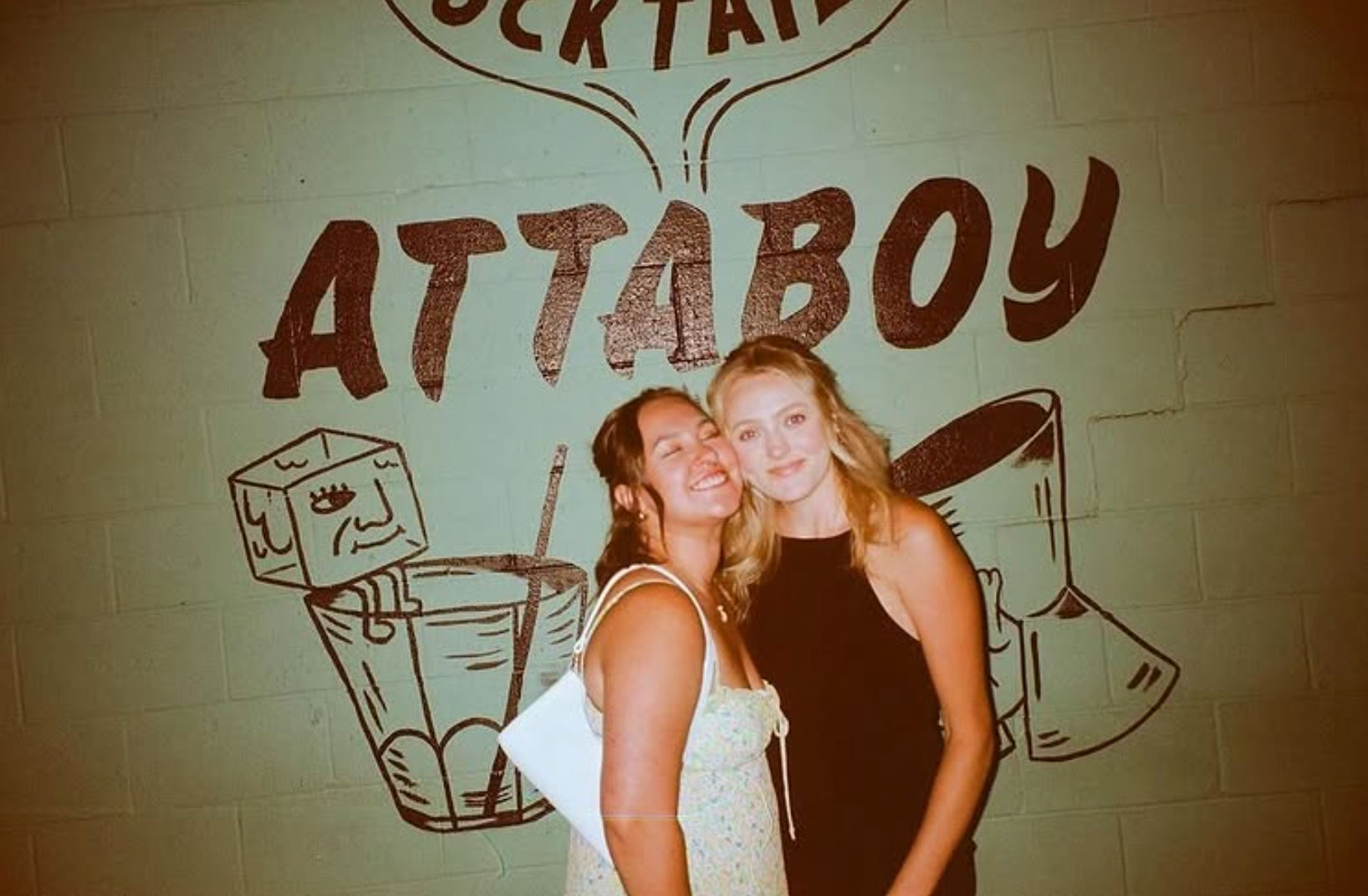 Two friends smiling and posing together in front of a green brick wall with a large cocktail-themed mural and bold lettering, night-out bar vibe.